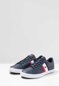Navy blue leather sneakers with white rubber soles, featuring red and white stripes and a green Lacoste logo on the side.