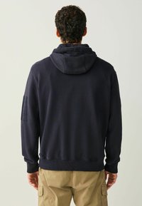 Tumesinine kapuutsiga dressipluus, millel on nööriga kapuuts, soonilised varrukasuu ja vöökohal. Vasakul varrukal on plaastriga detail. Pehme kanga tekstuur.