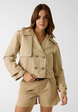 Kobieta nosząca beżową, krótko skrojoną, dwurzędową kurtkę oraz pasujące do niej szorty z wysokim stanem i brązowym paskiem na jednolitym tle.