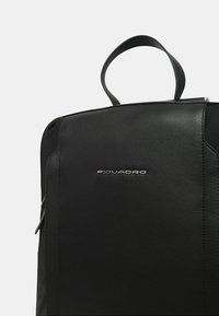 Zaino in pelle nera con texture liscia, dotato di manico superiore e dettagli del logo discreti. Design semplice, forma rettangolare, senza hardware visibile.