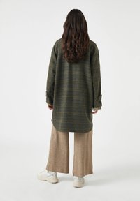 Over-sized grønternet frakke med knappede manchetter, parret med beige brede bukser og hvide sneakers, mod en simpel hvid baggrund.