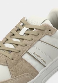 White and beige sneaker featuring suede and synthetic materials, with flat laces, "TOMMY JEANS" branding, and a textured sole.