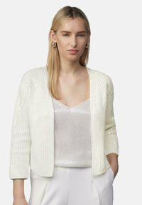 Weißer Pailletten-Cardigan mit gerippter Textur, offenem Vorderdesign und Dreiviertelärmeln, getragen über einem glänzenden, hellen Oberteil.