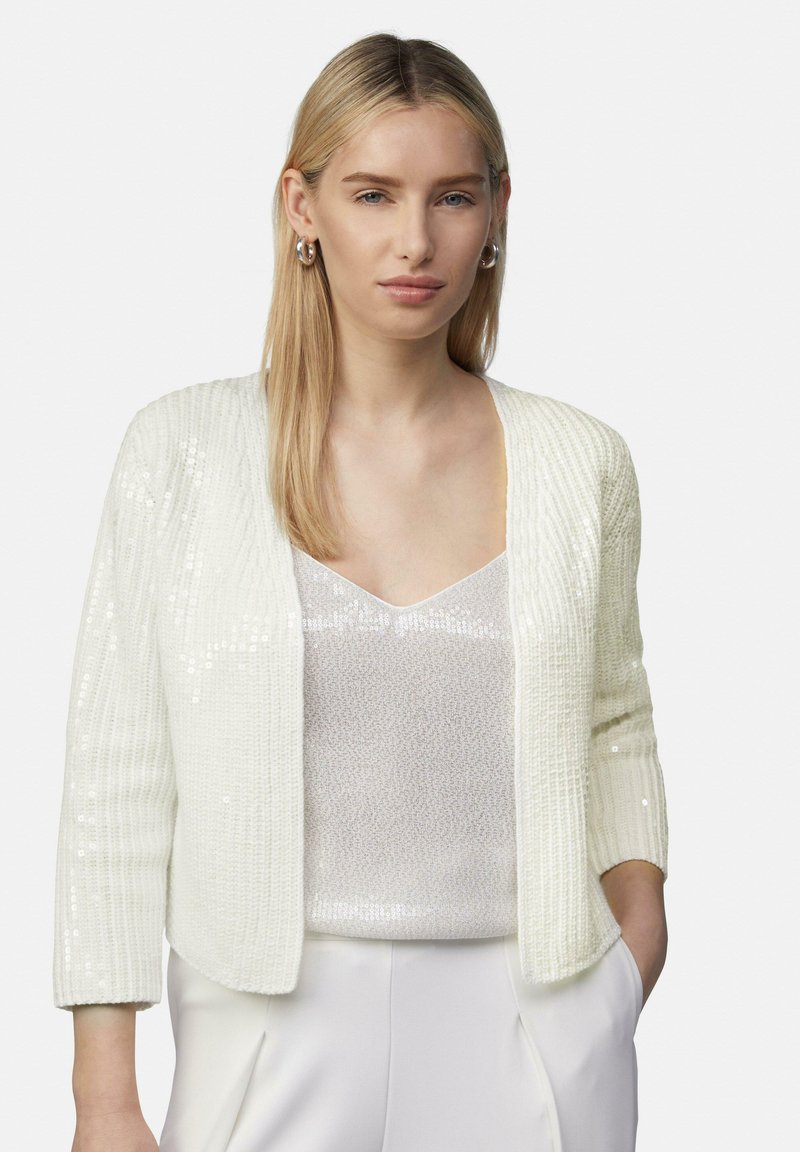 Weißer Pailletten-Cardigan mit gerippter Textur, offenem Vorderdesign und Dreiviertelärmeln, getragen über einem glänzenden, hellen Oberteil.