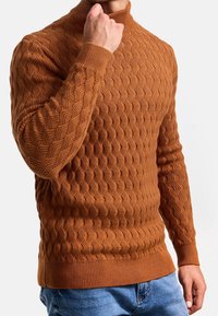 Homme portant un pull en maille texturée couleur rouille avec un col montant, la main touchant le menton, vêtu d'un jean bleu sur un fond uni.