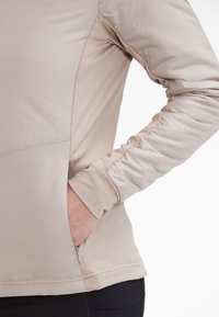 Lys beige atletisk jakke med en glat tekstur, der har sammensnørede ærmer, en sidelomme og et sømløst design.