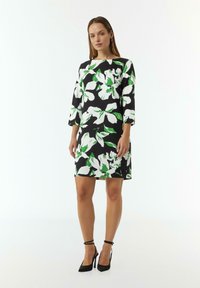 Schwarzes Kleid mit weißem und grünem Blumenmuster, Dreiviertelärmel, knielang, kombiniert mit schwarzen Riemchen-High-Heels.