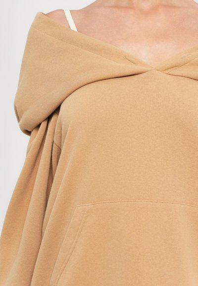 Femme portant un sweat-shirt beige à épaules nues avec une bretelle de soutien-gorge blanche visible sur son épaule droite.