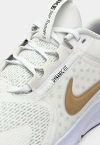 Scarpa sportiva bianca con tomaia in rete, dettagli grigi, spesse stringhe bianche e un logo Nike swoosh dorato. Presenta un'etichetta "Dynamic Fit" sulla linguetta.