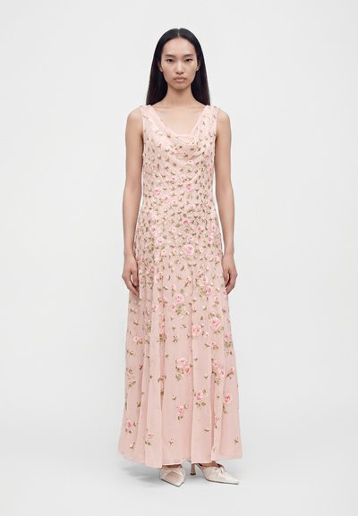 Femme aux cheveux noirs et lisses portant une robe rose sans manches, longue jusqu'au sol, avec une broderie florale et des chaussures en satin pointues.