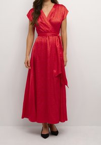 Kvinna som bär en ljusrosa omlottklänning med V-halsringning, kappryttar, knytmidja och flytande kjol, tillsammans med svarta spetsiga skor.