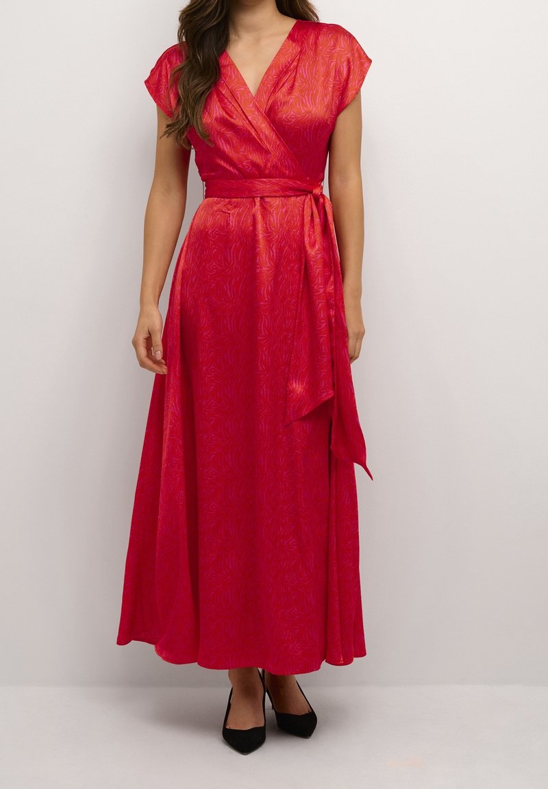 Kvinna som bär en ljusrosa omlottklänning med V-halsringning, kappryttar, knytmidja och flytande kjol, tillsammans med svarta spetsiga skor.