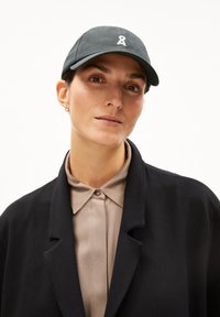 Dark green baseball cap with a white embroidered ribbon logo. The wearer also has a beige collared shirt and a black jacket.