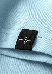 Hellblauer Stoff mit gerippter Textur, der an der Naht ein schwarzes Etikett mit einem weißen Herzschlag-Logo aufweist.