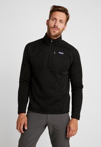 Schwarzer Fleece-Pullover mit einem Viertelreißverschluss und hohem Kragen, ausgestattet mit einem kleinen lila Patagonia-Logo auf der Brust. Glatte Textur.