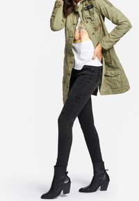 Olivgrön jacka med fickor, vit t-shirt med grafiskt tryck, svarta skinny jeans och svarta klackboots med ribbad textur.