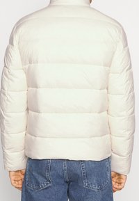 Person, der eine hellbeige Steppjacke und blaue Jeans trägt, von hinten gezeigt, vor einem einfarbigen, hellen Hintergrund stehend.
