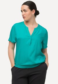 Blusa teal a maniche corte con scollatura a V, realizzata in tessuto liscio. Presenta cuciture sottili e una vestibilità rilassata, abbinata a pantaloni neri.