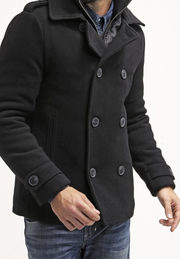 Homme portant un manteau noir à double boutonnage, superposé sur une chemise bleue et un jean, sur un fond blanc uni.