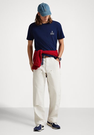 Μπλε ναυτικό t-shirt με λογότυπο, εκρού παντελόνι με φαρδιά μπατζάκια, κόκκινο πουλόβερ δεμένο στη μέση, ανοιχτό μπλε καπέλο και μπλε αθλητικά παπούτσια με λευκές λεπτομέρειες.