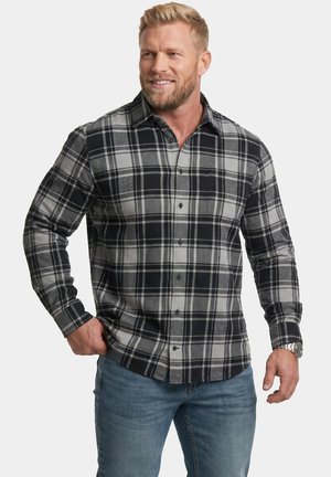 Lächelnder Mann, der ein schwarz-grau kariertes Hemd mit Knöpfen und blaue Jeans trägt, steht mit einer Hand in der Tasche vor einem einfarbigen Hintergrund.