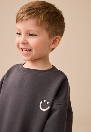 Junger Junge lächelt zur Seite, trägt einen dunkelgrauen Sweatshirt mit einem kleinen weißen Smiley-Gesicht auf der Brust vor einem beigen Hintergrund.