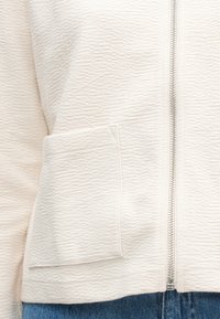 Helles beiges Reißverschluss-Jacke mit texturierter Stoffstruktur. Verfügt über eine Seitentasche und ein minimalistisches Design. Getragen zu blauen Jeans.