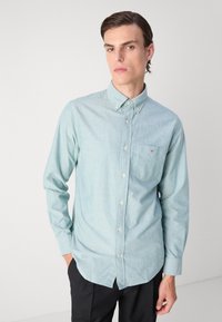 Camicia button-up azzurro chiaro realizzata in un tessuto testurizzato, con una tasca sul petto e un piccolo logo sulla tasca. Maniche lunghe e colletto classico.