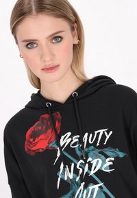 Sudadera negra con un gráfico de una rosa roja y el texto "BELLEZA DE ADENTRO HACIA AFUERA". Cuerdas ajustables y detalles plateados en la capucha.