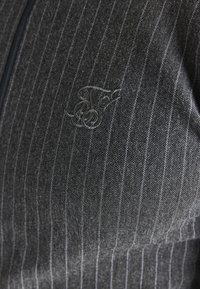 Grå jacka med dragkedja, med en nålrandig mönster, mjuk textur och broderad logotyp i en kontrasterande färg på bröstet.