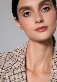 Złoty naszyjnik z małym, przezroczystym kryształowym wisiorkiem, noszony na tle strukturalnego beżowego żakietu. Tkanina ma wplecione wzory z ciemnymi i metalicznymi akcentami.