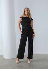 Top nero scollato abbinato a pantaloni ampi a vita alta. Texture liscia con minimi dettagli. Modella che indossa sandali con tacco chiari.