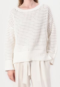 Woman wearing a white loosely knit sweater with diamond-shaped holes and ribbed cuffs, paired with cream high-waisted pants with a belt.