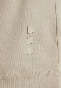 Beige Stoff mit gerippter Textur, mit drei kleinen quadratischen Laschen, die vertikal ausgerichtet sind. Sichtbare Nähte am Rand.