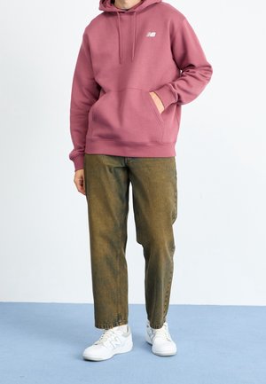 Persoon draagt een mauve New Balance hoodie, olijfgroene broek en witte New Balance sneakers, staand op een blauwe vloer tegen een witte muur.