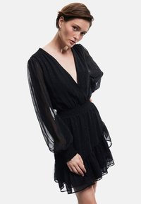 Schwarzes Kleid aus transparenter Stoff mit langen Puffärmeln, V-Ausschnitt, elastischem Taillenbund und geschichteten Rüschen am Saum. Mit subtilen Mustern.