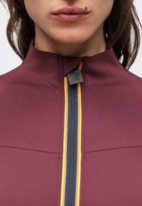 Vinröd jacka med hög krage och frontdragkedja med svart beslag samt gula, blå och orange ränder längs dragkedjan. Slät yta.