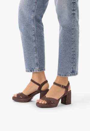 Piedi con sandali in camoscio marrone con tacco a blocco e punta aperta, con cinturini alla caviglia, abbinati a jeans azzurri corti.