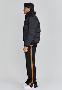 Schwarze, puffige Jacke mit hohem Kragen, kombiniert mit schwarzen Hosen, die einen braunen Streifen und Druckknöpfe an den Seiten haben, ergänzt durch weiße Sneaker.