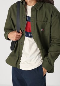 Olivově zelená bunda na zapínání s kapsou na prsou, v kombinaci s krémovým tričkem s výraznou červeno-modrou grafikou. Jsou vidět tmavé kalhoty.