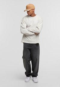Camisa de manga larga color blanco sucio, jeans negros holgados y zapatillas blancas. El modelo lleva una gorra color beige con visera curvada. Diseño simple y casual.