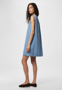 Kvinne med krøllete hår iført ermeløs lys blå denimkjole og brune flate sko, stående i profil mot en ensfarget bakgrunn.