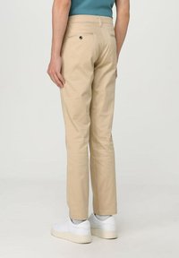 Pantaloni beige in cotone con vestibilità dritta, tasca posteriore visibile con dettaglio a bottone, indossati con sneaker bianche, mostrati da dietro.