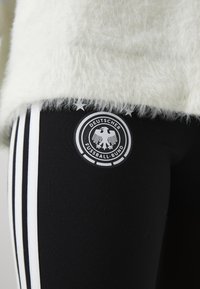 Leggings pretas com três riscas brancas e um logótipo circular com uma águia e "Deutscher Fussball-Bund", colocados numa parte superior branca e fofa.