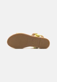 Sandalias amarillas con suela texturizada marrón, que cuentan con correas ajustables y cierre de hebilla. La parte inferior tiene un patrón circular y el logo de la marca.