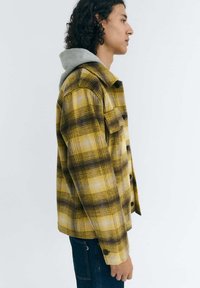 Gelb-braun karierte Jacke mit grauem Kapuzen, geknöpftem Vorderteil und Seitentaschen, im Profil vor einem hellen Hintergrund präsentiert.