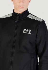 Chaqueta negra con cremallera y cuello alto, que presenta el logotipo blanco de EA7 y detalles a rayas en los hombros. Fabricada con un tejido suave y texturizado.