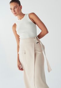 Bela rebrasta majica na naramenice uparena s visokim bež hlačama koje imaju širok pojas sa srebrnom kopčom i bočne džepove. Glatka tekstura tkanine.