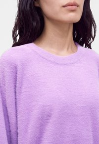 Personne aux longs cheveux foncés portant un pull lavande doux et duveteux avec un col rond sur un fond clair.