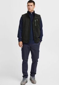 Czarna bezrękawnik nałożona na granatowy sweter, z zamykaną na zamek kieszenią na piersi, noszona z bojówkami i szarymi sneakersami. Prosty, funkcjonalny design.